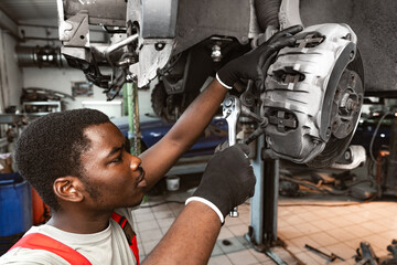 African male auto-mechanic repairing car brakes under the car in auto service - obrazy, fototapety, plakaty