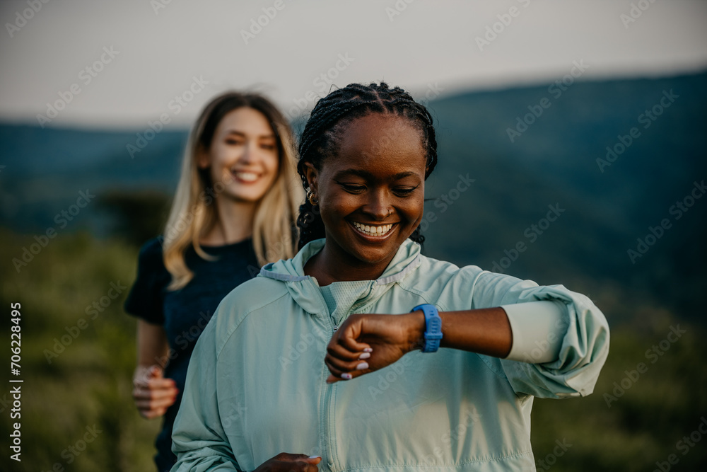 Wall mural black and white women jogging together, glancing at smartwatches for fitness updates