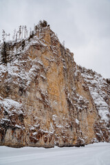 Fototapeta na wymiar High cliff rising above snow-covered river, winter landscape, rocky cliff, deserted place in the forest, winter journey to the mountains.