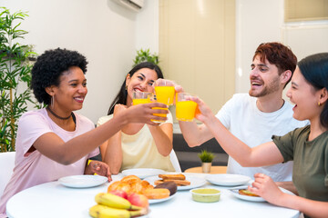 Multi-ethnic friends toasting with orange juice having breakfast at home - obrazy, fototapety, plakaty