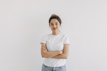Asian Thai woman wear white with bun hair, smiling, crossing arms over chest, looking at camera and standing over white background wall.