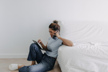 Asian Thai woman holding and talking on phone while using on writing on tablet, busy work sitting on floor and leaning sofa in room apartment alone in day time.