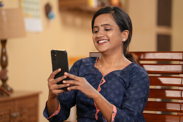 Happy young Indian girl busy using mobile phone at home - concept of social media sharing,...