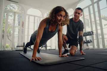 Multiracial trainer with young woman in sports clothing doing push-up exercises at the gym - obrazy, fototapety, plakaty