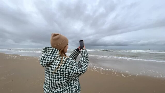 a girl stands on the ocean shore and films nature on her phone. Stormy clouds, high contrast. filming in 4K
