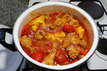 Stew chicken legs with tomato sauce, pineapple chunks, carrots, and tomatoes in an enameled pot on a gas stove.

