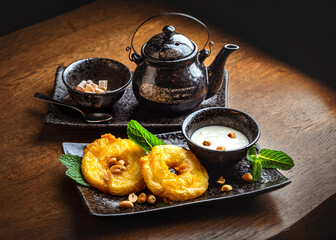 Asian sweets and tea. Fried pineapple slices in batter, coconut milk sauce, ceramic teapot and a bowl with cane sugar.