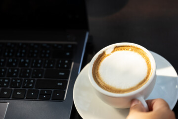 Business women hand holding white latte coffee cup and laptop on black table background.Hot coffee cup with laptop on desk when work from home.