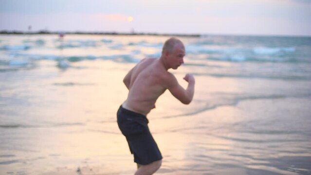 Young muscular man practicing boxing exercises at the sea beach. Male sportsman is practiced self defense alone near ocean shore. Training of martial arts outdoor at sunset. Slow motion Close up