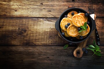 Delicious cottage cheese pancakes or syrniki with fresh blueberry in cast-iron pan on dark wooden...