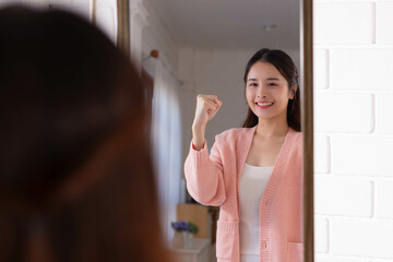Smiling beautiful asian woman looking at herself in the bathroom mirror at home. - obrazy, fototapety, plakaty