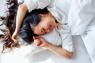 Attractive happy Asian woman lies on a white linen bed in the morning. The young girl smiles and...