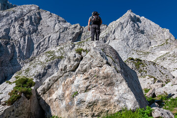 Hiker man stands on massive rock formation along hiking trail from Fusine Lake to Mangart saddle in Tarvisio, Julian Alps, Friuli Venezia Giulia, Italy, Europe. Rugged extreme alpine terrain in summer