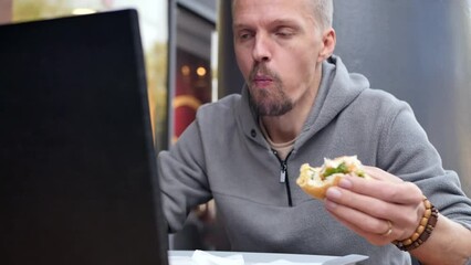 A hungry handsome man eats a hamburger and at the same time discusses a discussion while working online via video conference using a laptop. a quick fast food snack during a busy work schedule