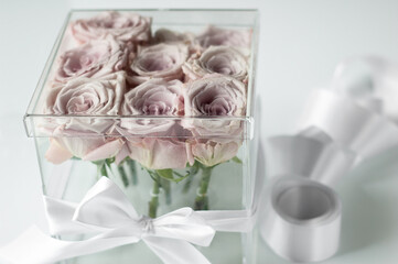 Light pink roses in a clear transparent flower box on a white table. Square glass gift box.