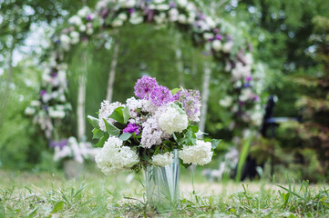 A bouquet of lilac flowers, purple giant onions, and other flowers in a small bucket as a decorative element against the background of a wedding arch. - 706327846
