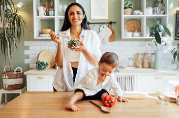A happy vegan family in the kitchen at home. A young attractive mother and her little son are eating vegetables. A smiling woman with a plate of salad and her cute baby.