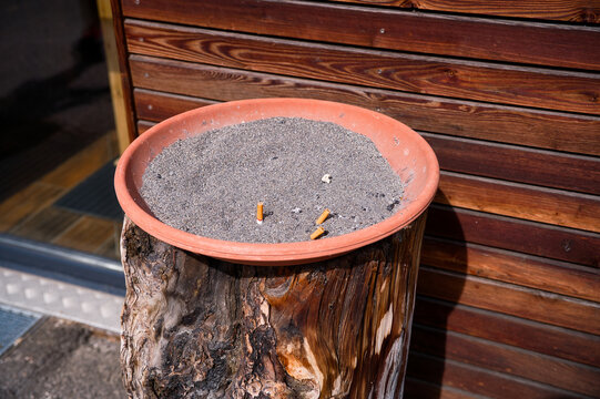 Eine mit Sand gefüllte Tonschale auf einem Baumstumpf an einer Holzwand mit Zigarettenstummeln im Sand