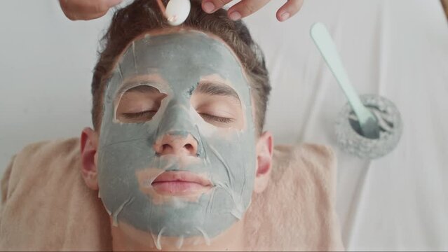 Top down view of young man of mixed race lying on couch with eyes closed while getting beauty procedure with jade roller and masks on face at spa studio