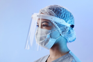 Medium close up with side view of serious woman doctor in medical face shield, mask and sterile hat looking away standing at pastel blue background - Powered by Adobe