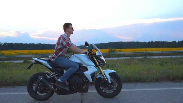 Young man riding on a modern sport motorbike. Handsome motorcyclist in a shirt and glasses driving his motorcycle on country road with sunflowers field at background. Slow motion Side view Close up