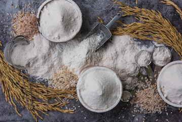 Rice flour Thai organic in a measuring spoon with wooden bowl  on a pile of white rice, ear of paddy and  strainer on concrete background, Top view flat lay, Food and baking ingredient.