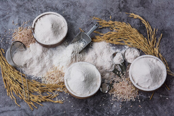 Rice flour Thai organic in a measuring spoon with wooden bowl  on a pile of white rice, ear of paddy and  strainer on concrete background, Top view flat lay, Food and baking ingredient.