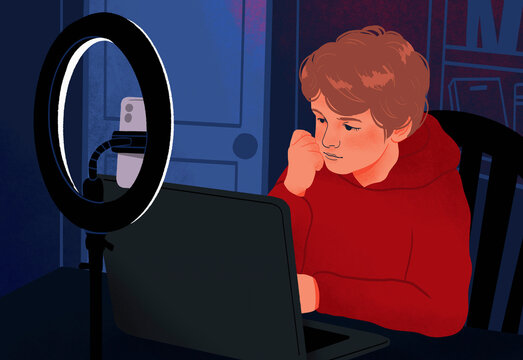 Boy using laptop in dark bedroom behind ring light and smart phone
