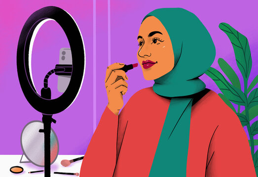 Female influencer in hijab giving makeup tutorial with lipstick behind ring light and smart phone

