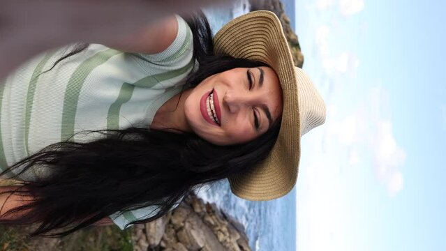 Vertical video referring to the mobile screen of a woman making a video call with the beach in the background, greeting her friends with her smartphone
