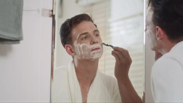 Medium close up of young man using razor while shaving beard in front of mirror with shaving foam applied on face