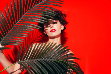 Fashion beauty portrait of young confident brunette woman with evening stylish makeup and perfect clean skin. Sexy model with curly hair posing in studio. With red bright lips. Tropical palm leaf