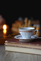Vintage wooden chessboard, books, glasses, pen, cup of tea or coffee and scented candle on the table. Dark academia concept. Selective focus.