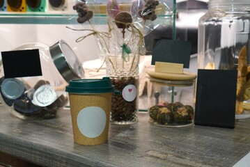a paper cup for a coffee drink on the bar next to glass jars with coffee beans and green tea with sweets on a stick