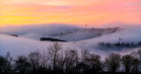 Panoramic view of foggy winter scenery in Sauerland Germany. Fluffy low mist clouds in the land of 1000 hills and valleys. Evening twilight after sunset with colorful sky gradient above the horizon.