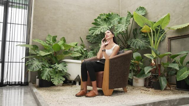 Modern young woman talking on the phone, working remotely or relaxing in the solitude of a minimalist architectural concrete style summer exotic plant foliage garden with tropical plants. Blithe