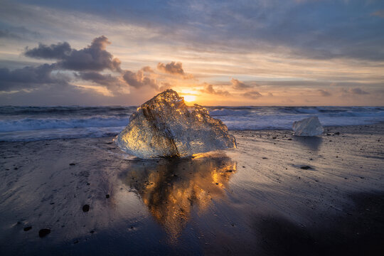 Reflection of an Ice fragment on black sand beach illuminated orange by a setting sun in Iceland