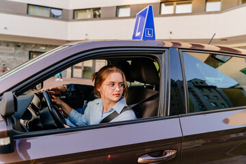 Young woman learning how to drive car together with her instructor. Driving school. Happy driving student. Brown car with blue L plate on a roof.