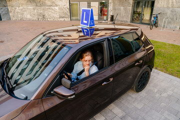 Brown car with blue L plate on a roof. Above view of thoughtful young female with seat belt. Caucasian Teen Learning Driver's Education. The girl failed her driving test.