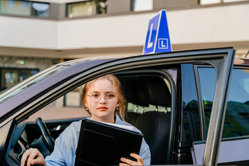Student driver class, learning how to drive. Portrait of young female taking lessons seriously looks into the camera gets out of the car from the driver's seat holding clipboard.