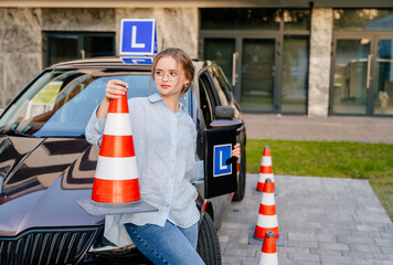 The girl poses near a learner's car with an L sign on the roof. She holds a traffic cone and a folder with the letter L in her hand. The concept of learning driving skills in a driving school.
