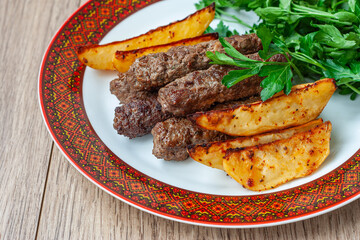 Lula kebabs on a white plate with a red border with baked potatoes and greens.