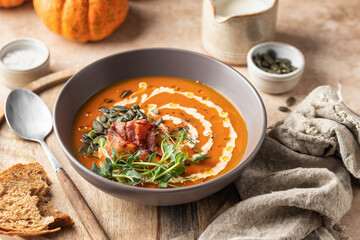 Pumpkin cream soup decorated with cream, pumpkin seeds, bacon, microgreens and olive oil on brown...