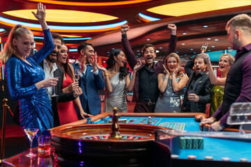 Attractive Multiethnic People Taking Risks and Placing Bets on a Roulette Wheel in a Casino. Croupier Focused on Table with Tokens, while Ball Spins on the Ball Track. Player Celebrates a Winning Bet - obrazy, fototapety, plakaty