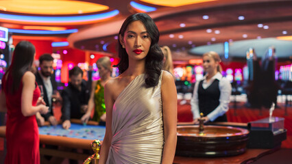 Advertising Campaign Portrait of a Sophisticated Asian Woman in a Stunning Dress Posing and Looking at Camera in a Glamorous Casino with Hustlers and Gamblers Playing Roulette in Vibrant Background