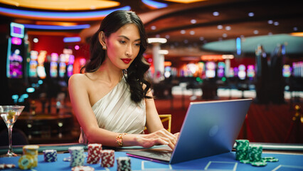 Glamorous Asian Woman at Casino Table, Engaged in Online Betting Games on Laptop, After Winning Bet...