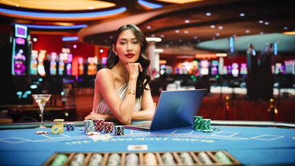 Glamorous Asian Woman at Casino Table, Engaged in Online Betting Games on Laptop, After Winning Bet...