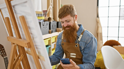 Confident redhead young man, a smiling artist, carefully drawing on smartphone in art studio...