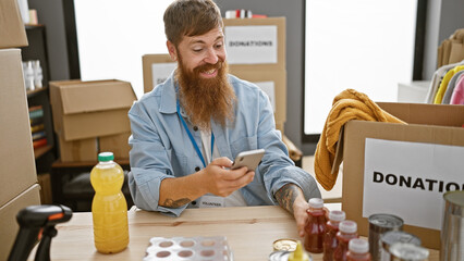 Confident young redhead man volunteering with joy at charity center, smiling while typing on smartphone sitting at table