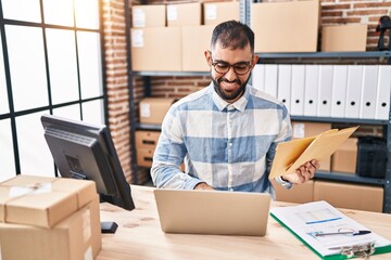 Young hispanic man ecommerce business worker using laptop holding packages at office